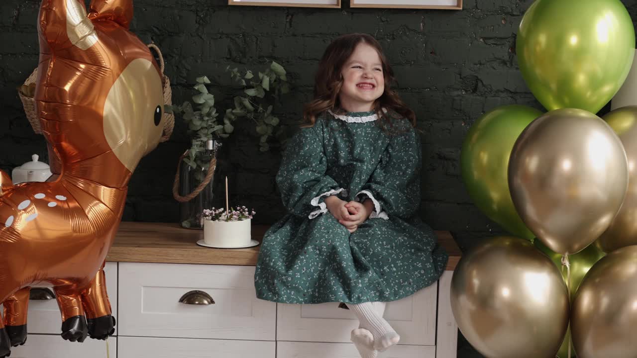 孩子的生日聚会。可爱快乐的小女孩穿着节日时尚的衣服坐在绿色砖墙背景的马桶上，房间里装饰着气球，鹿形气球和带蜡烛的生日蛋糕视频素材