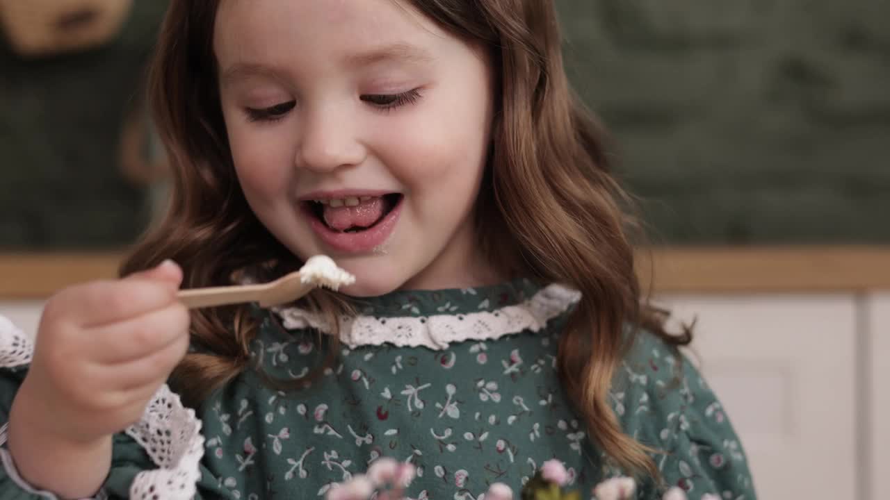 生日聚会。快乐可爱的小女孩4-5岁，穿着节日服装，在时尚的家庭厨房里愉快地吃着生态木勺，她装饰的鲜花便当和白色奶油生日蛋糕视频素材