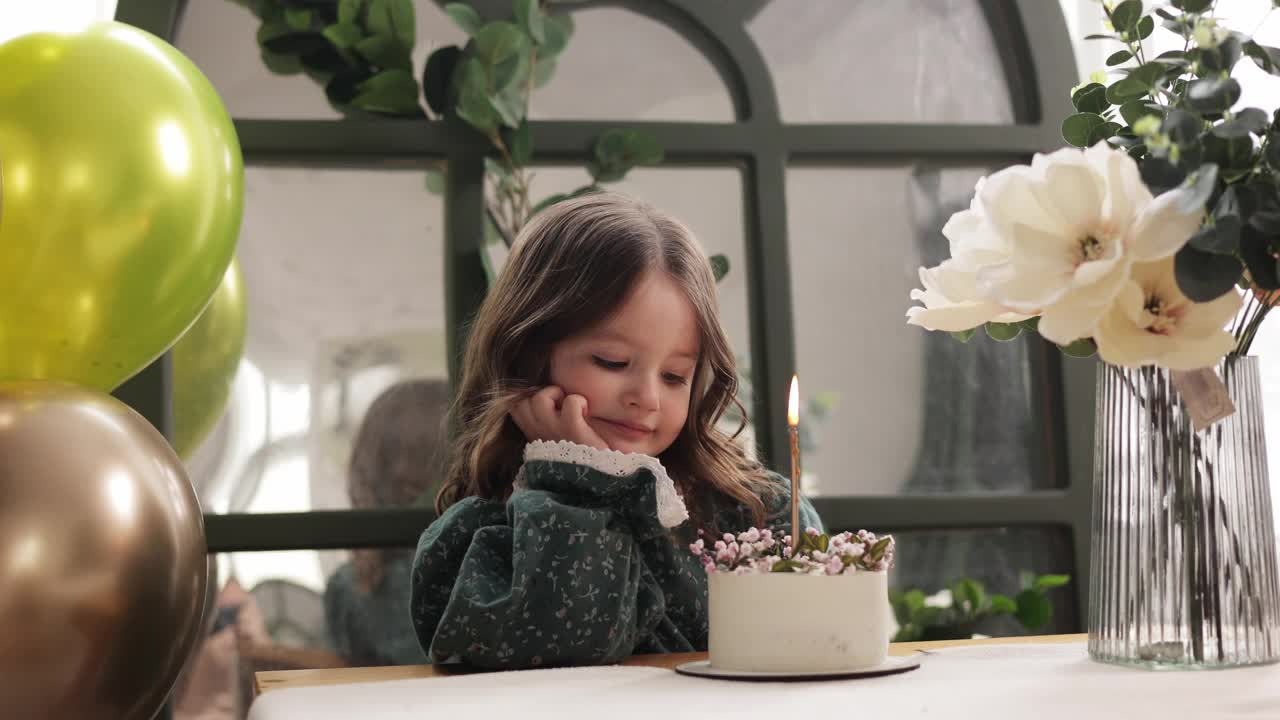 孩子的生日聚会。可爱的小女孩坐在桌子旁，用生日蛋糕装饰鲜花和点燃蜡烛，许愿。穿着节日服装的孩子在房间里拿着气球，花瓶前放着一束花视频素材