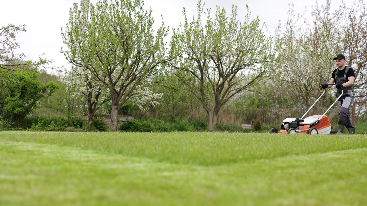身穿防护服的专业园丁正在后院用现代汽油无绳割草机割绿草。季节性景观设计工作。背景是盛开的树木视频素材