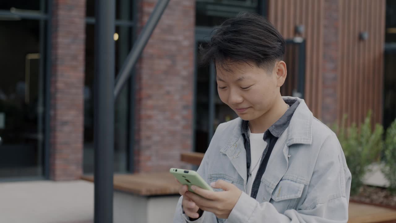 快乐的亚洲学生用智能手机走在现代城市街道上的慢动作视频素材