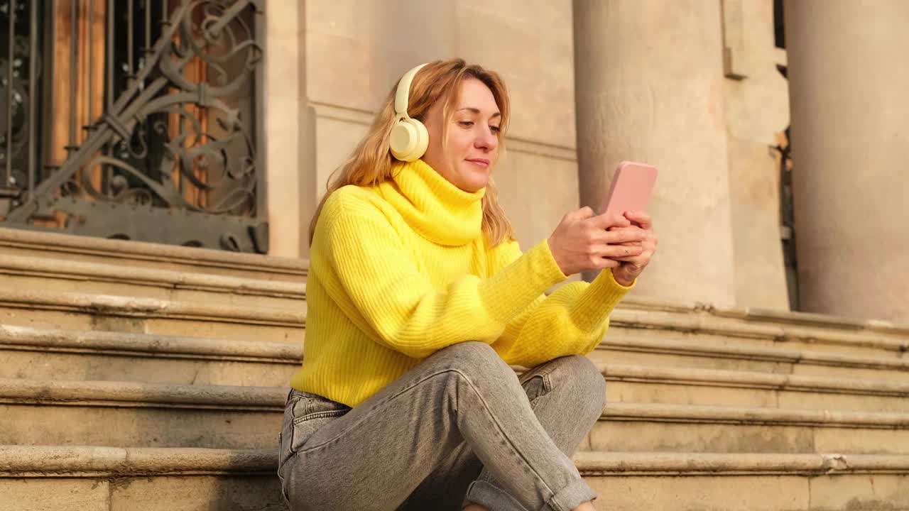 戴着耳机的女人坐在外面的楼梯上，微笑着看着手机。成熟的成人学生观念。城市的生活方式视频素材