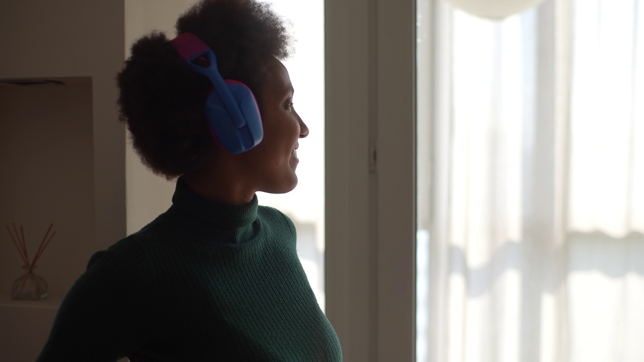 一个无忧无虑的非洲裔美国女人，戴着耳机，独自在家窗边跳舞。微笑的黑人女性听着音乐感觉自由视频素材