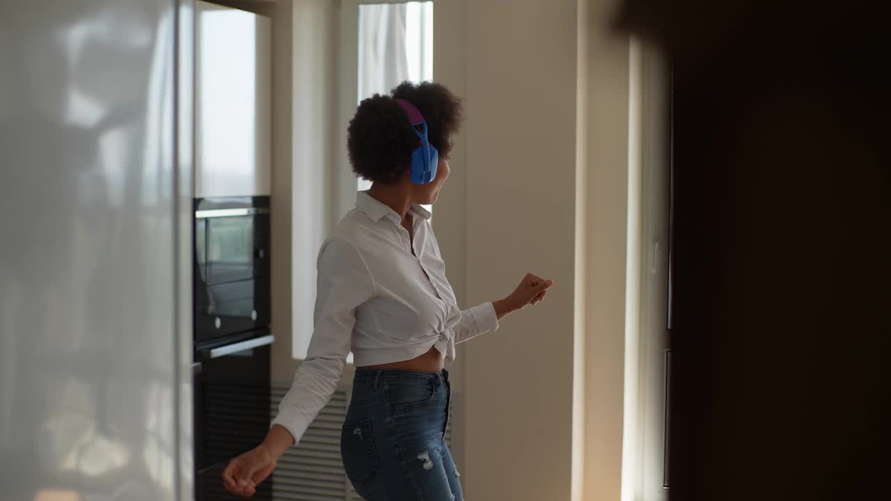 一个戴着耳机，闭着眼睛跳舞唱歌的快乐的非洲裔年轻女子，在现代公寓内部举手。视频素材