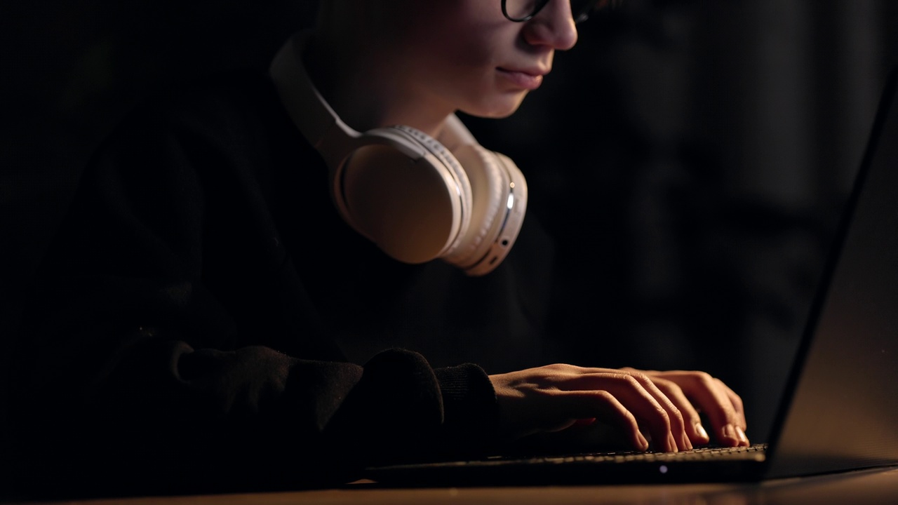 年轻的电脑玩家戴着眼镜，脖子上戴着耳机，在笔记本电脑键盘上打字，在黑暗的房间里玩电脑游戏，电子竞技。学生正在学习或在个人电脑上的社交媒体交流。视频素材