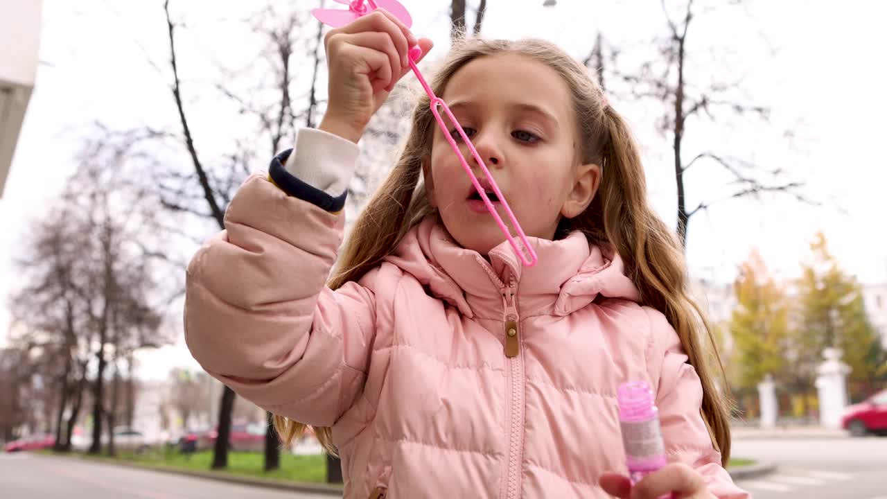 穿着粉色夹克的小女孩在街上吹肥皂泡秋天，冬天，春天视频下载