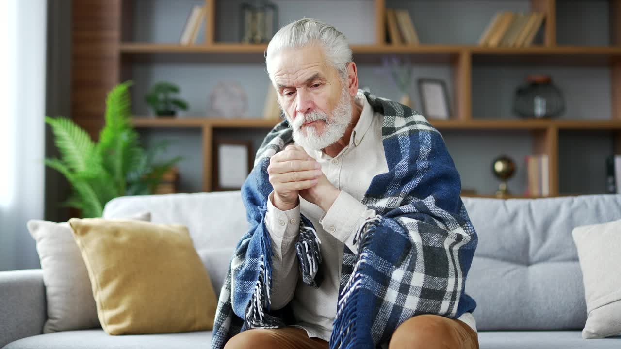 一位白发苍苍的老人裹着毯子坐在家里客厅的沙发上冻住了。心烦意乱的成熟退休男冷室内。视频素材