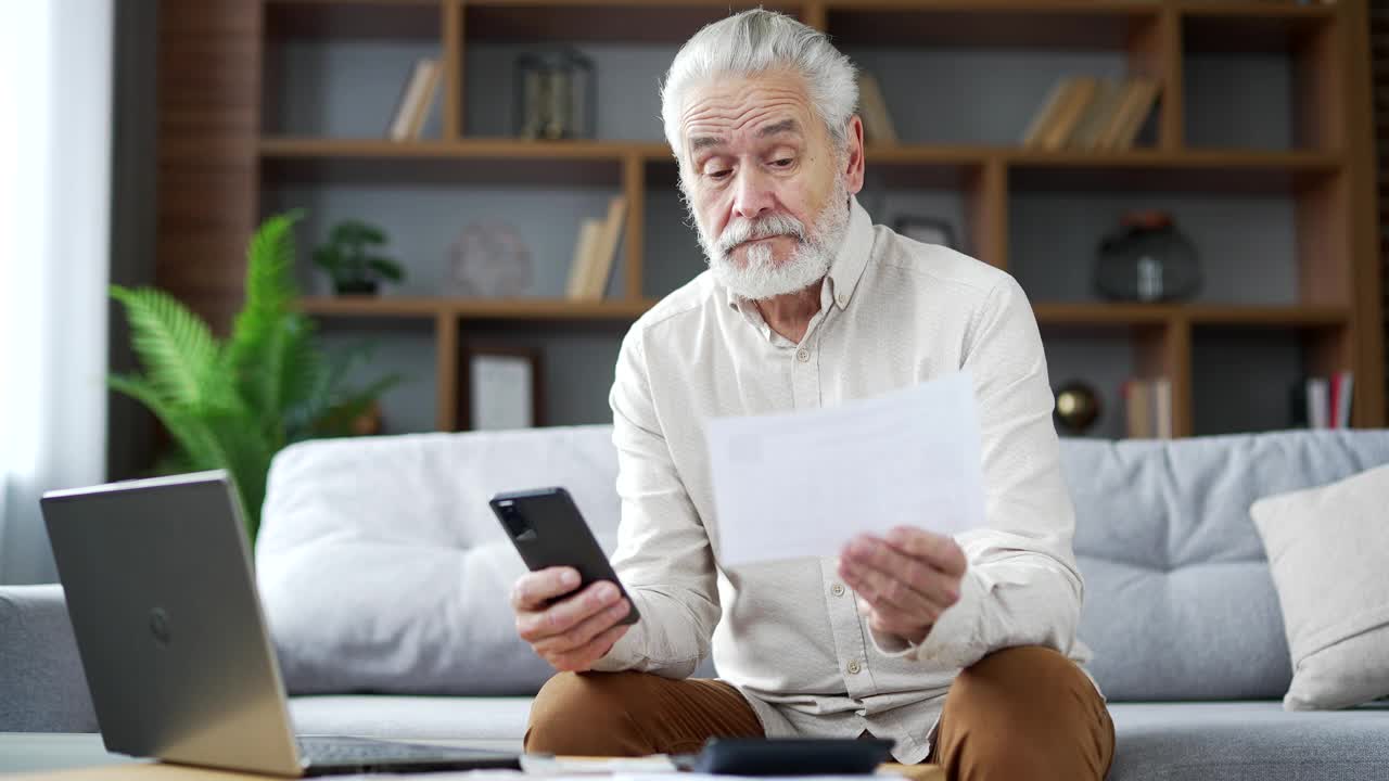 老人坐在家里的沙发上用智能手机计算家庭财务有困难。视频素材