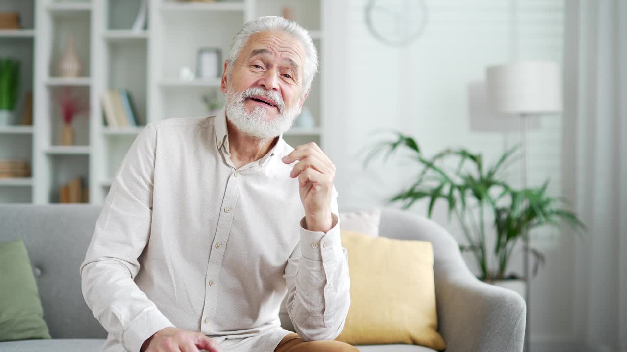 摄像头的观点。开朗的白发老人坐在家里客厅的沙发上看着摄像头正在进行视频通话视频素材