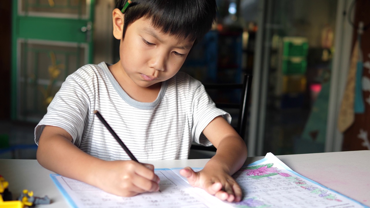 亚洲小男孩坐在课桌前写练习本。可爱的小学生在家里做作业，脸上带着微笑和专注。教育理念，学习，家校。视频素材