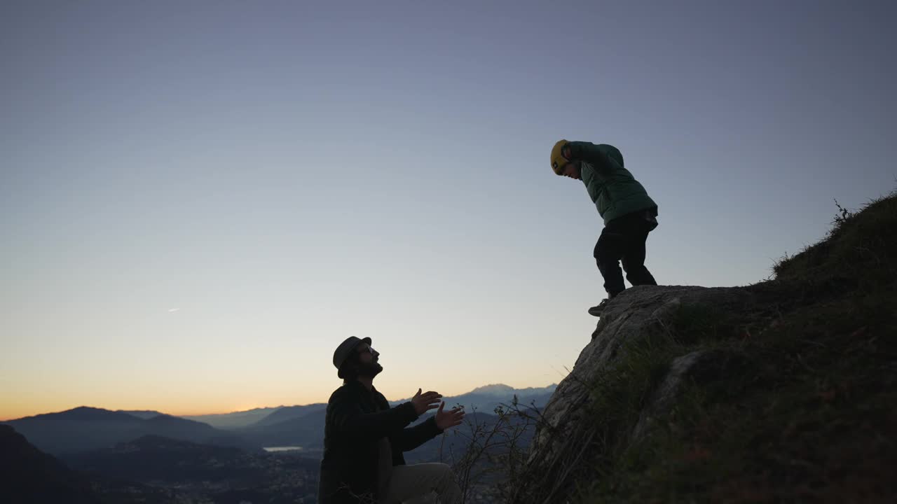 孩子从岩石上跳下来，投入父亲的怀抱，夕阳映衬出他的剪影视频素材