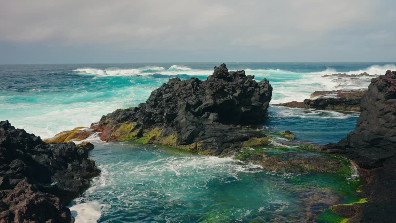 波涛汹涌的海浪冲击着布满火山岩的海岸线的慢镜头。蓝绿色的海水拍打着海岸。晴天，晴空万里。视频素材