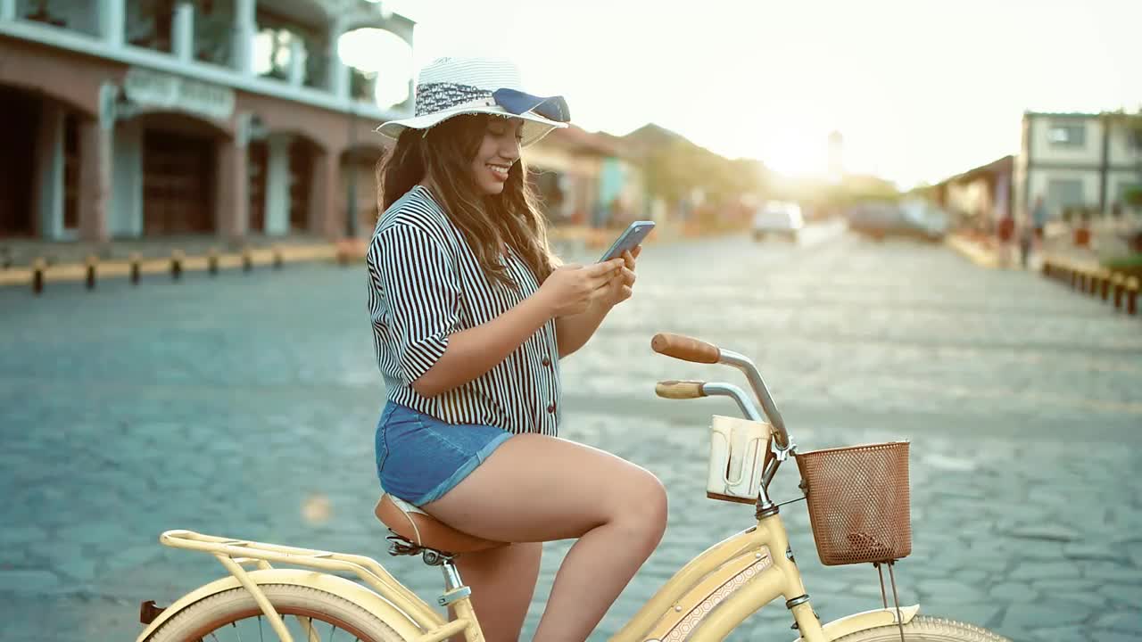 快乐的旅游女孩戴着帽子，骑着自行车，在尼加拉瓜格拉纳达的街上发短信。微笑着的旅游女孩骑着自行车在电话里发短信视频素材