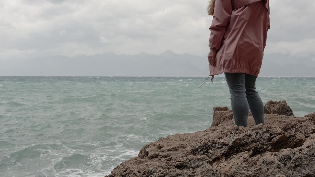 一个穿着粉色雨衣的女人站在海边的悬崖边上，看着暴风雨。缓慢的运动。视频素材