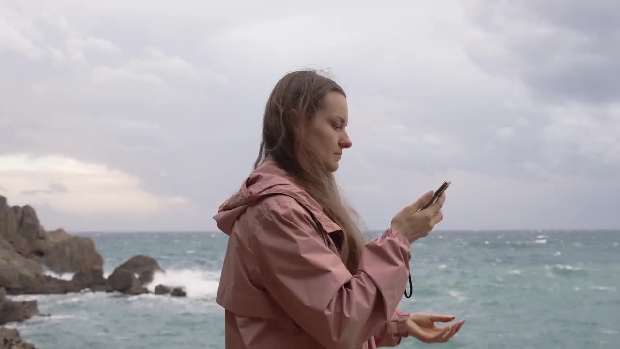 暴风雨中的年轻女子站在海边的岩石上，看着她手机上的东西。阴天，她穿着一件粉红色的雨衣。视频素材