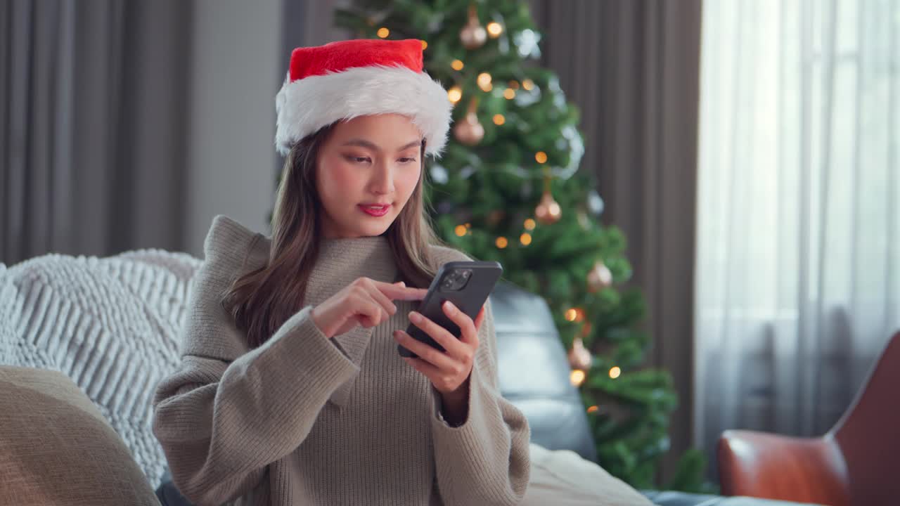 兴奋的亚洲女人戴着圣诞老人的帽子，用手机坐在家里圣诞树旁边的沙发上，庆祝成功，做出“是”的手势，收到好消息，购物打折，阅读留言视频素材