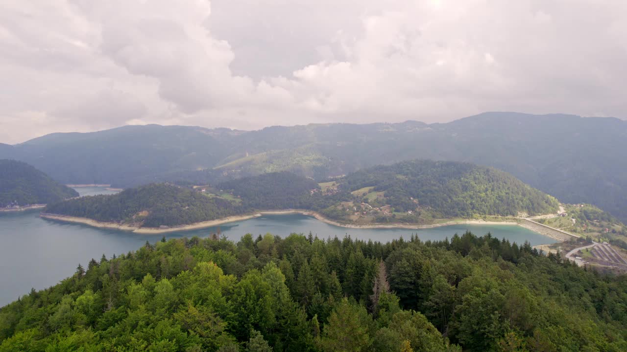 在多云的天气里，鸟瞰图中的蓝色湖泊周围树木繁茂的山丘视频素材