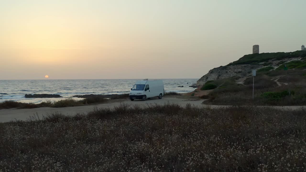 在意大利撒丁岛的日落时分，一辆露营车在曼努角的海滩和大海附近的灯塔上的空中无人机视图视频素材