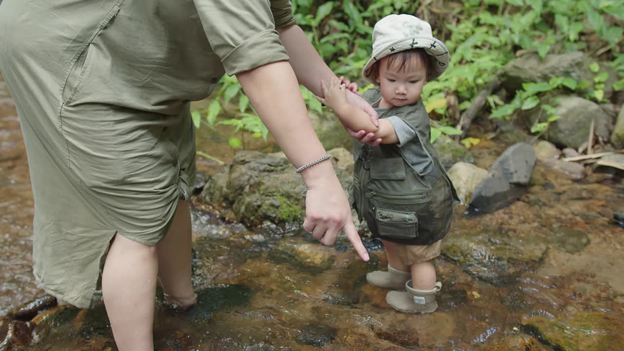 幸福的家庭妈妈牵着手可爱的小亚洲学步的女儿走在山河水溪在雨林中学习自然，探险探险者野营旅行之旅视频素材