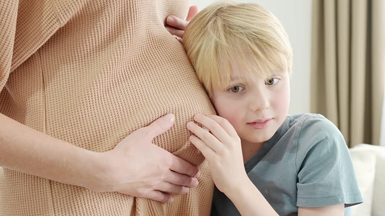 一个男生抱着他怀孕的妈妈，倾听着她的肚子。怀孕的母亲和她的大孩子在家里。未来的大哥。视频素材