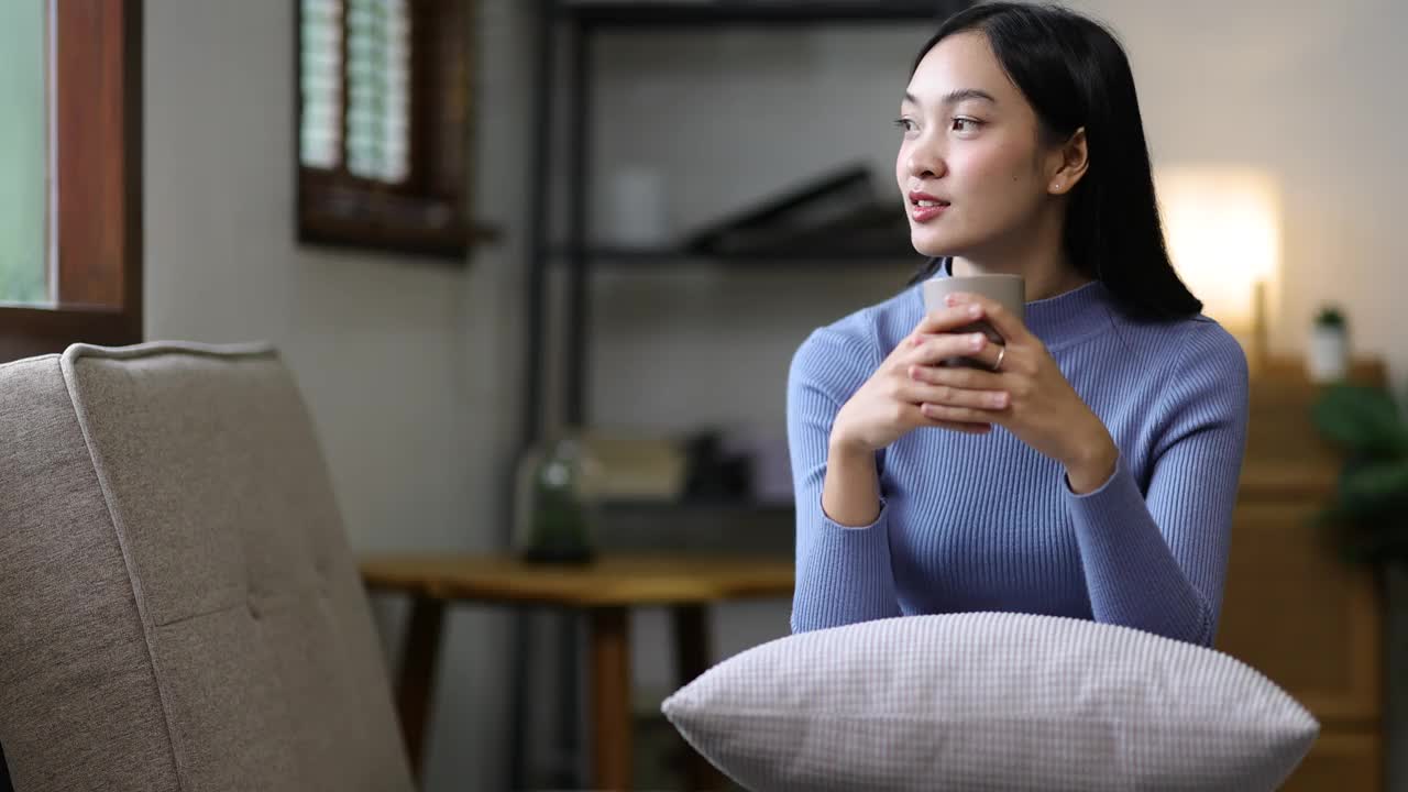 美丽的亚洲女人在家里一边喝咖啡一边在客厅的沙发上放松。视频素材