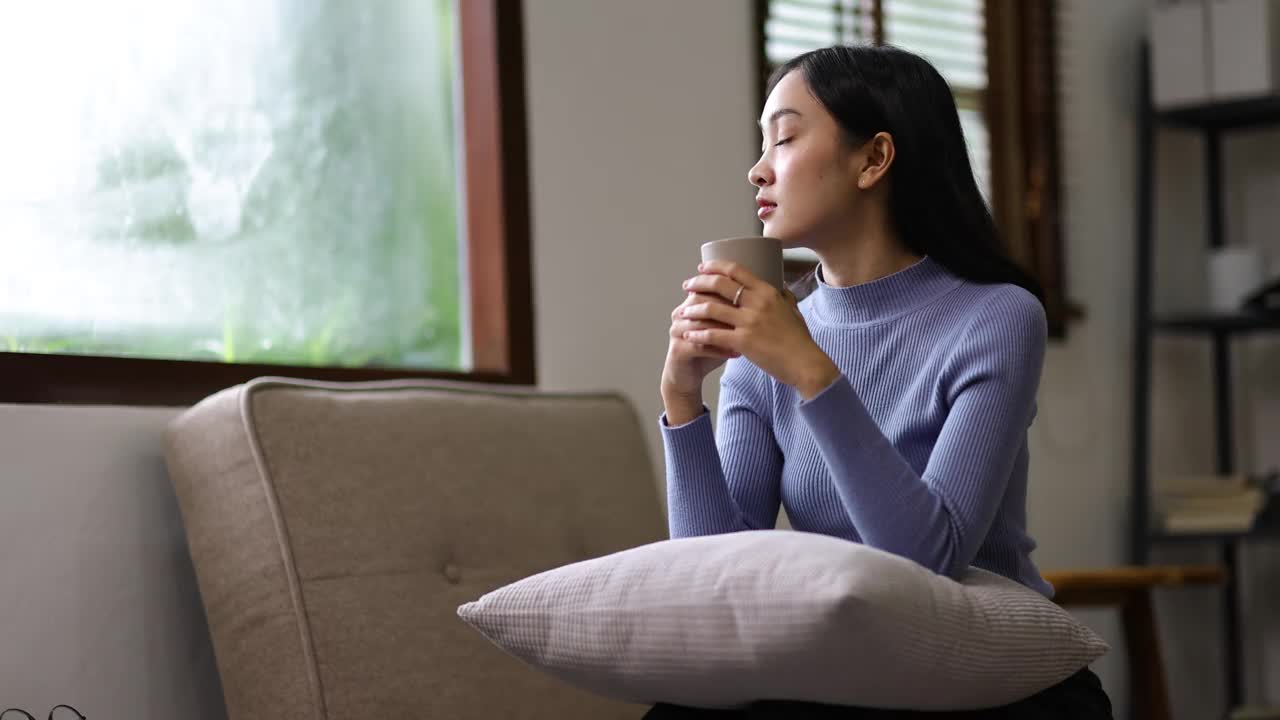 美丽的亚洲女人在家里一边喝咖啡一边在客厅的沙发上放松。视频素材