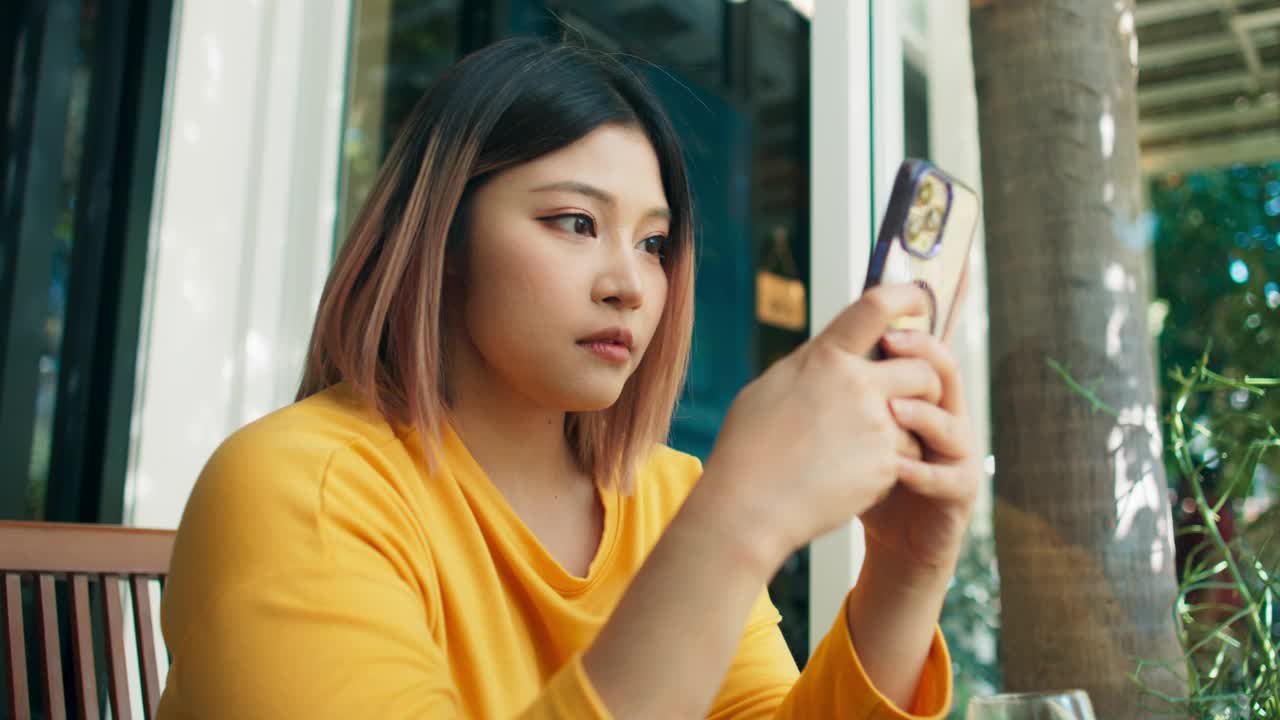 积极的年轻亚洲女性穿着休闲服装，在咖啡馆和餐厅的咖啡休息时间用手机与朋友聊天和上网，女性在闲暇时间放松社交媒体和互联网视频素材