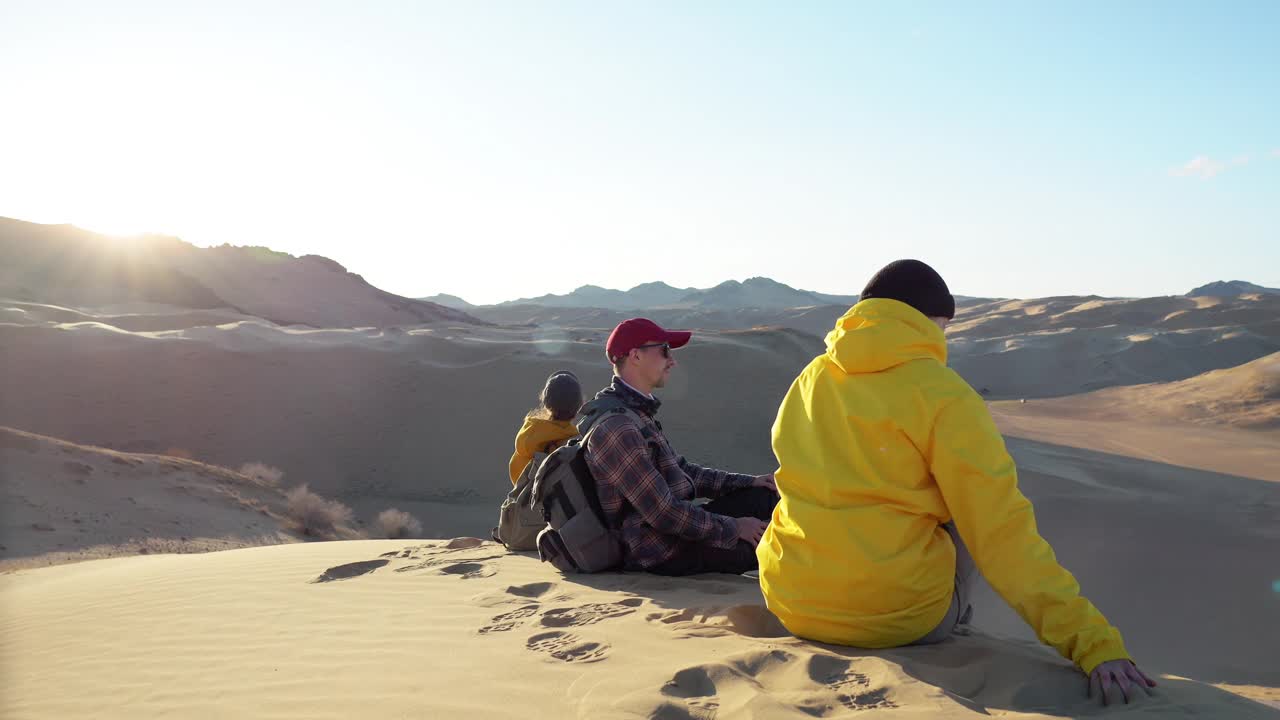 三个年轻的徒步旅行者一起在阳光下坐在沙漠的沙丘上放松视频素材