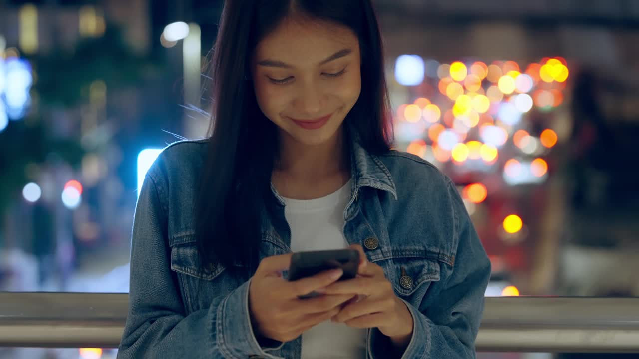 美丽的年轻亚洲女子站在城市街道在晚上使用手机。一个快乐的女人站在那里用手机打字视频素材