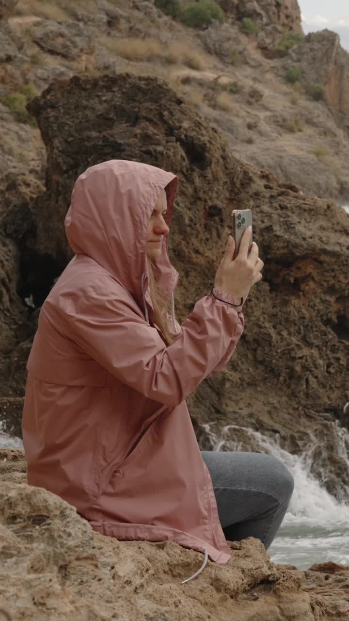 垂直视频。一个穿着雨衣的女人坐在尖锐的岩石上，用手机拍下了惊涛骇浪的大海。视频素材