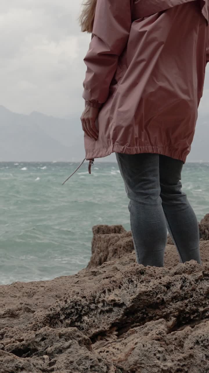 垂直视频。一个穿着粉色雨衣的女人站在海边的悬崖边上，看着暴风雨。缓慢的运动。视频素材