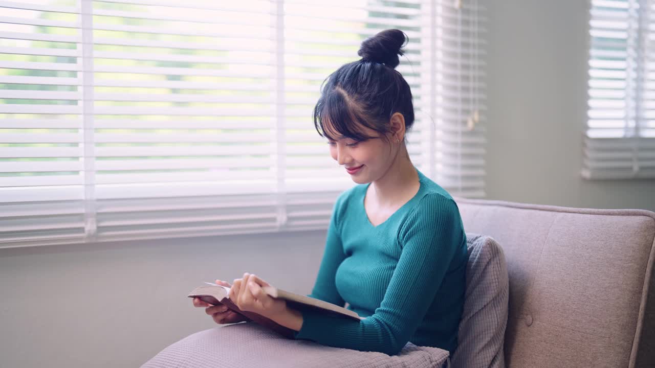 周末，一位亚洲年轻女子舒舒服服地坐在家里的沙发上看小说。当她沉浸在一本好书中，捕捉周末放松的宁静时刻。视频素材