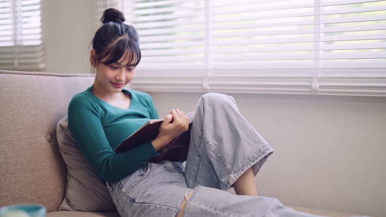 周末，一位亚洲年轻女子舒舒服服地坐在家里的沙发上看小说。当她沉浸在一本好书中，捕捉周末放松的宁静时刻。视频素材