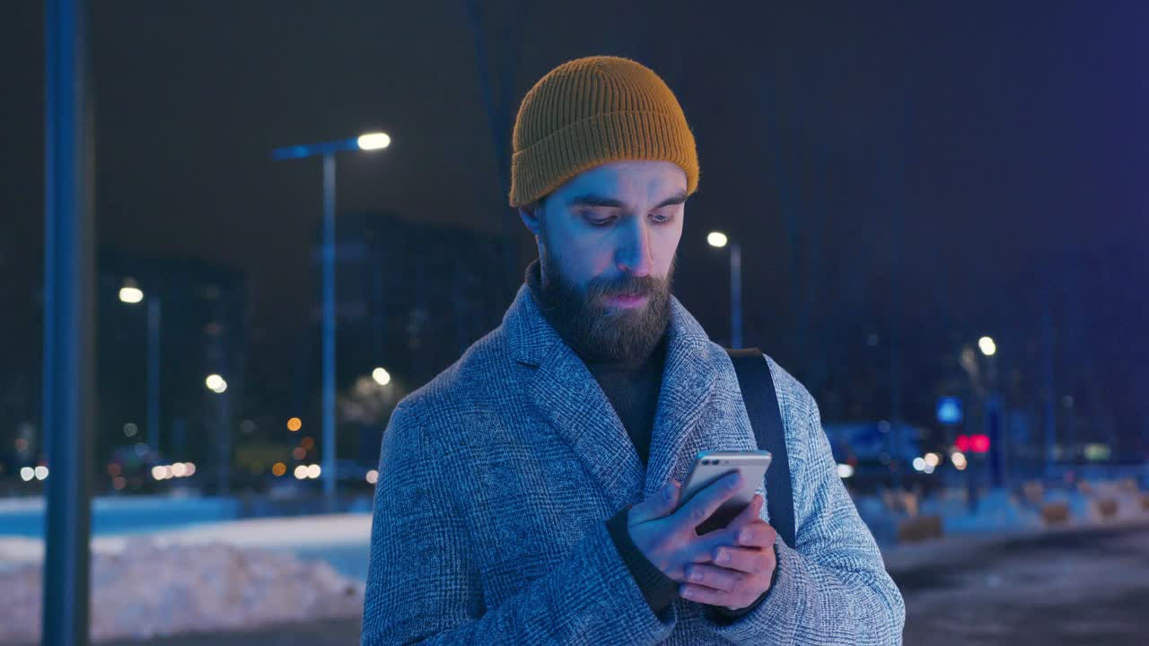 一位戴着黄色帽子，穿着保暖外套的中年男子在冬天的夜晚在城市的街道上使用智能手机视频下载