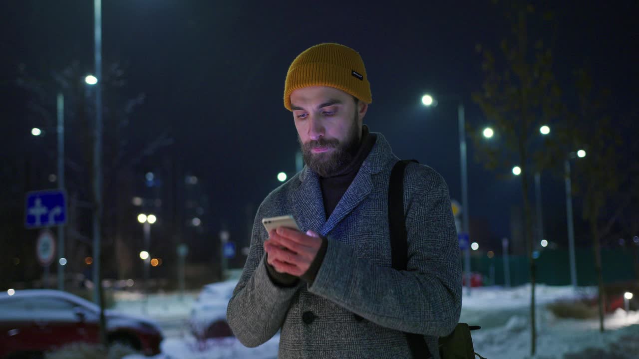 一位戴着黄色帽子，穿着保暖外套的中年男子在冬天的夜晚在城市的街道上使用智能手机视频下载