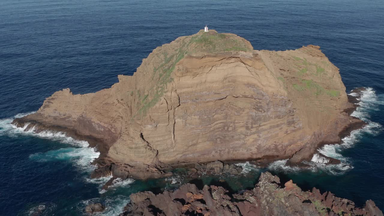 宁静的蓝色海洋中的巨石，日落时的灯塔。前景是火山岩，海浪滚滚而来。无人机绕岩石逆时针旋转。在葡萄牙马德拉岛的莫尼兹港。视频素材