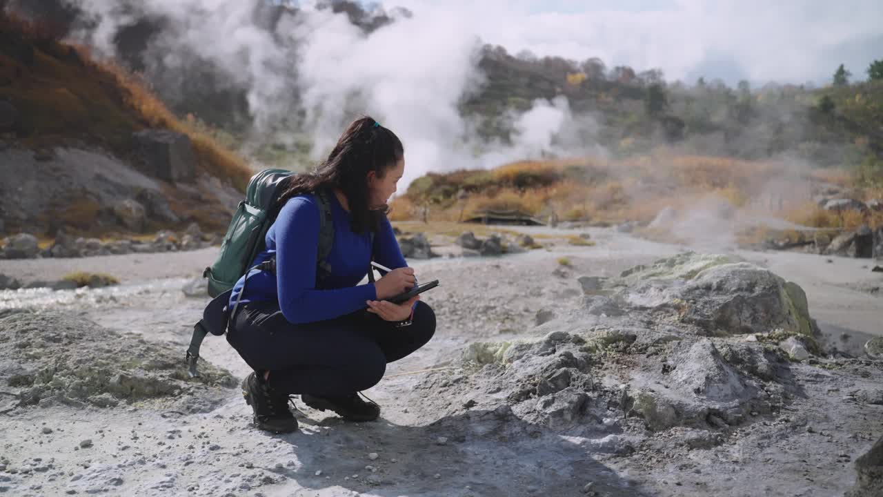 一位亚洲女性地质学家坐在热气腾腾的温泉附近的岩石区，观察着她的数字平板电脑上收集的数据，她的研究重点是地热能视频素材