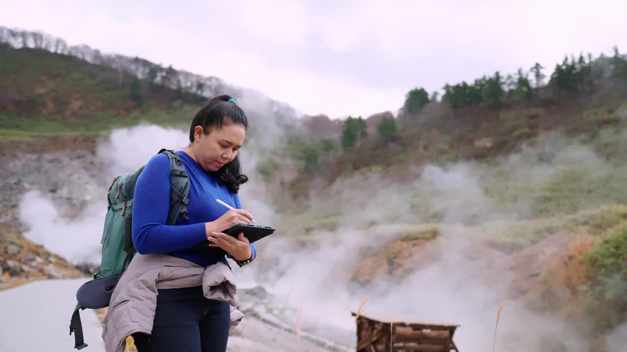 一个阴天，一位亚洲女性环保人士在日本东北秋田市森林山丘附近研究温泉，在她的电子平板电脑上记录她的观察结果。视频素材