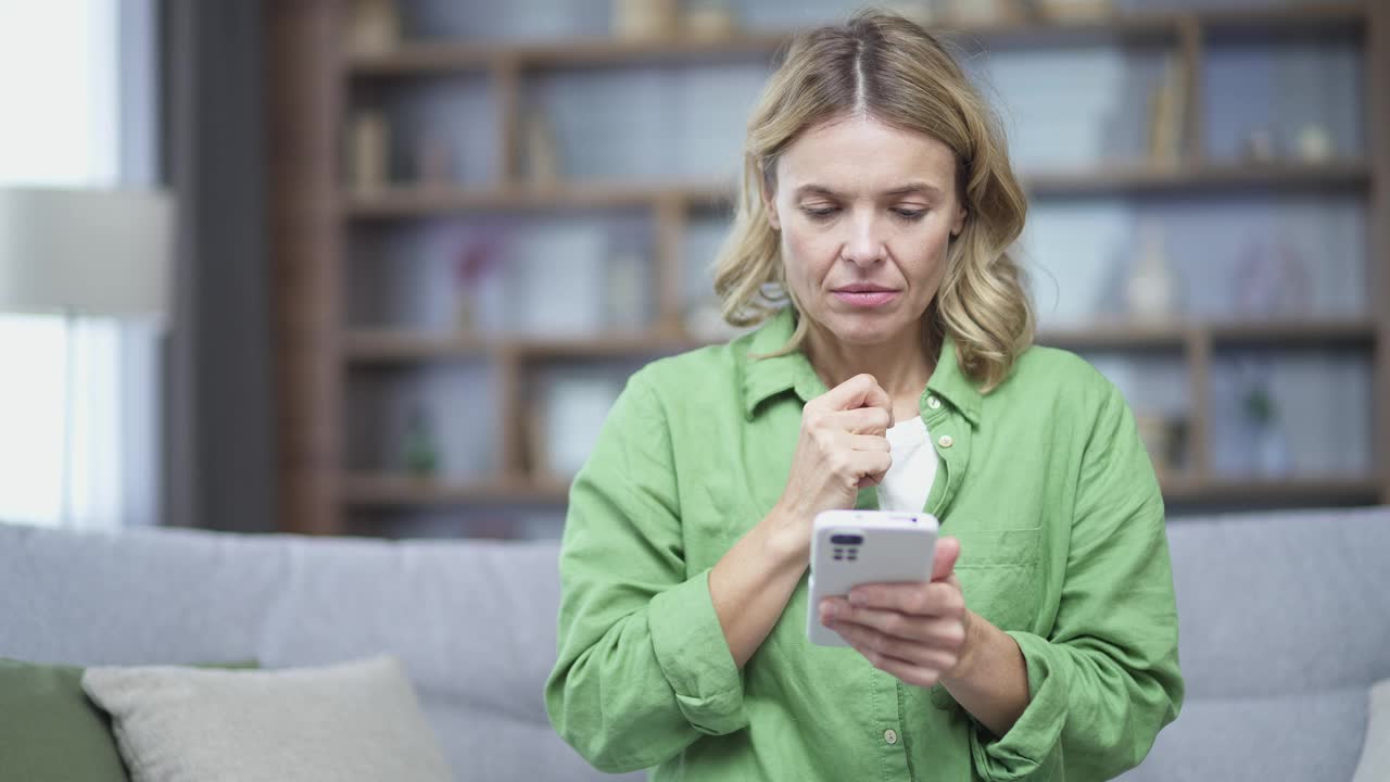 悲伤的金发成熟女人坐在家里的沙发上看智能手机上的坏消息。视频素材