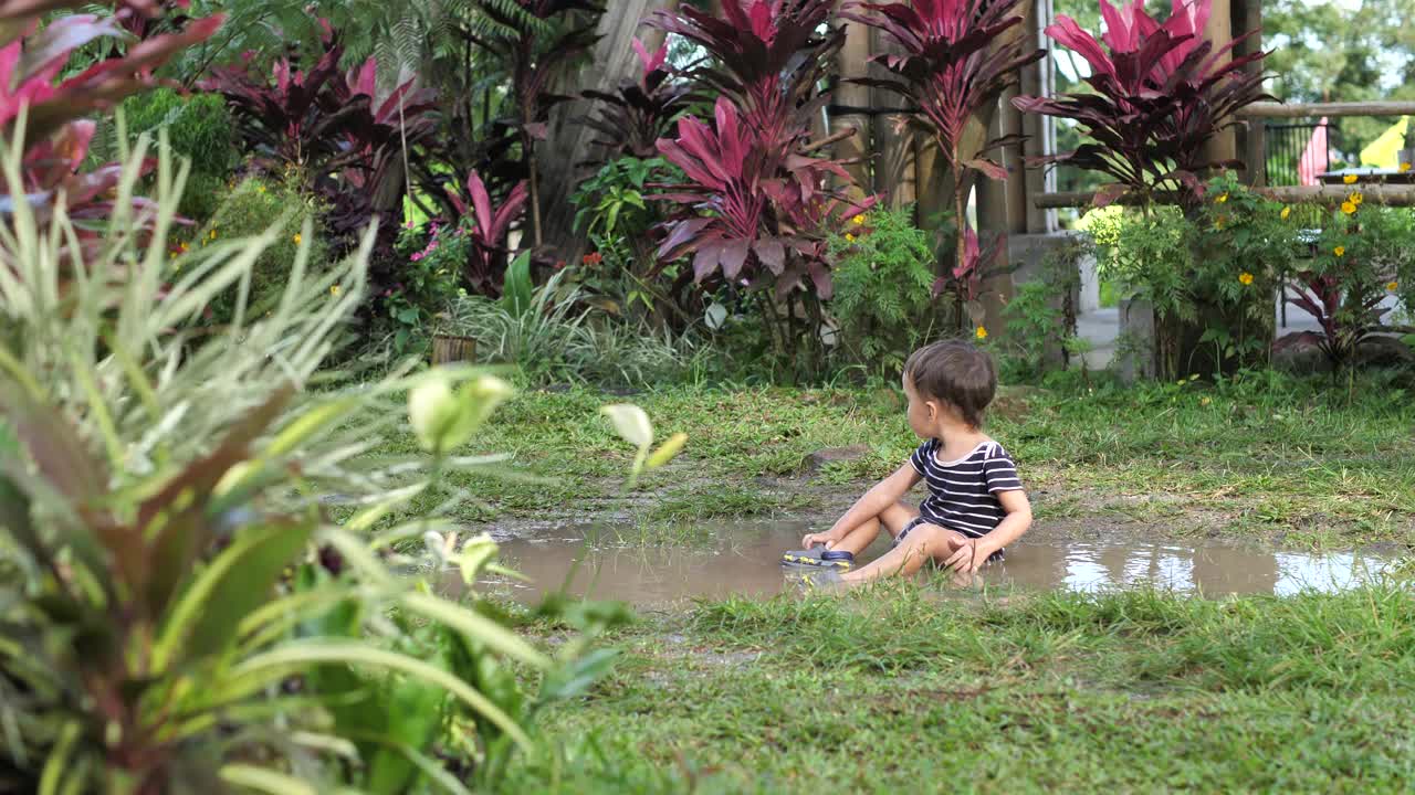 蹒跚学步的男孩坐在院子里的脏水坑里视频素材