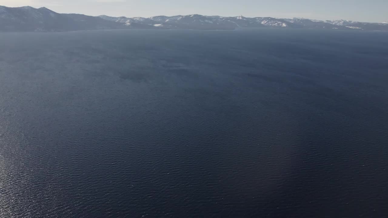 冬季，一架高空飞行的4K无人机在加州太浩湖上空拍摄。镜头慢慢向上平移，展现出远处白雪覆盖的内华达山脉，环绕着湖泊。视频素材