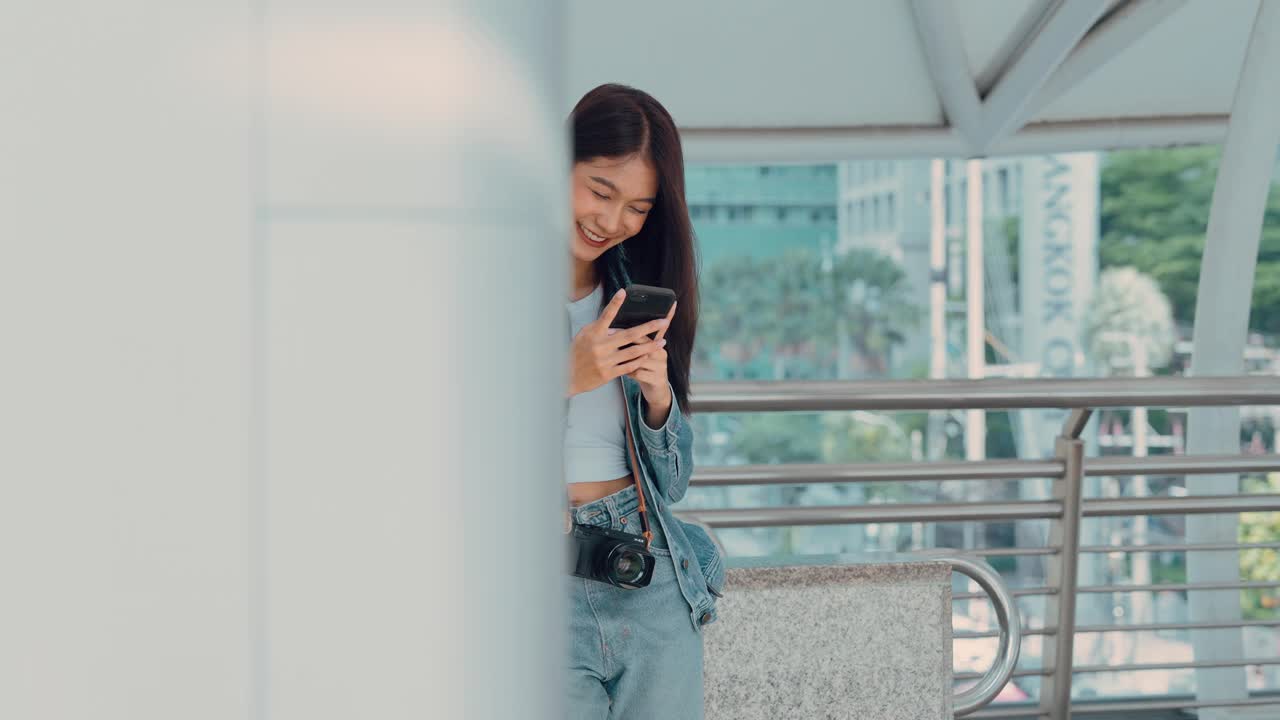 年轻的亚洲女子微笑着，在户外使用移动智能手机。穿着牛仔裤夹克拿着智能手机出现在公共城市的快乐美女。在线技术与传播理念视频素材