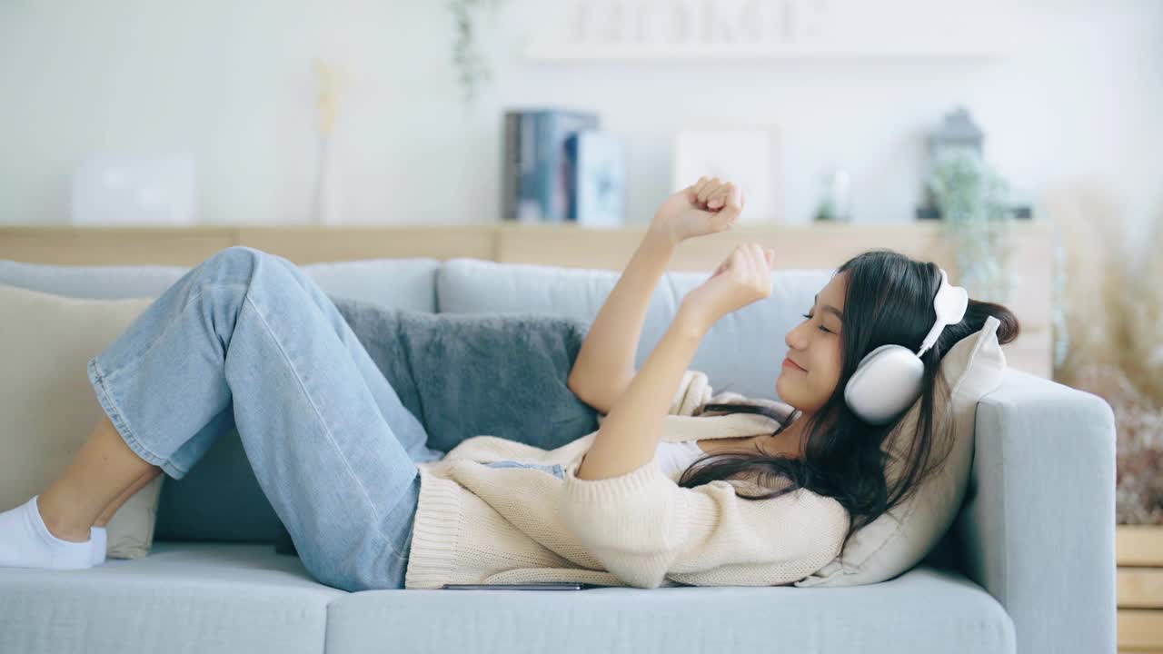 年轻的亚洲女子在家里客厅的沙发上听音乐。快乐亚洲女性使用手机，戴耳机，躺在沙发上视频素材