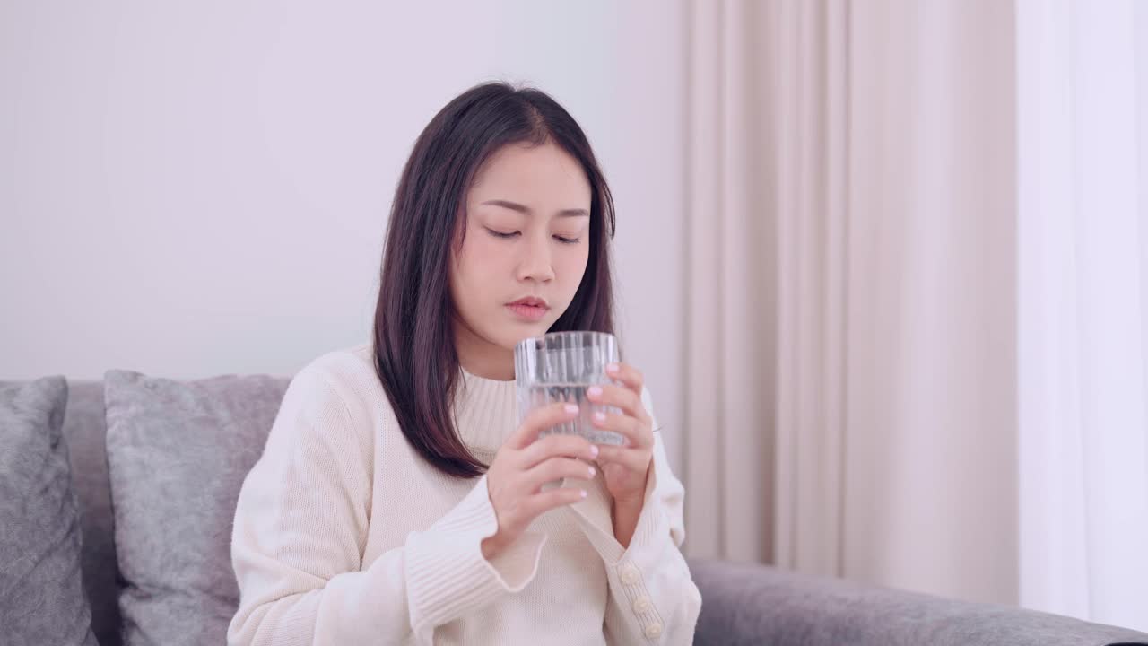 一名亚洲妇女坐在家里的沙发上喝水来缓解喉咙痛。一个自我照顾和安慰的时刻。视频素材