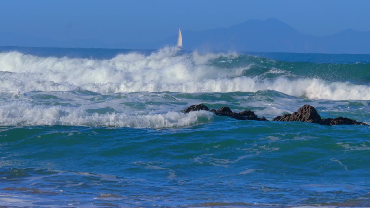 强烈的海浪拍打着海岸上的岩石，背景是一艘帆船视频下载