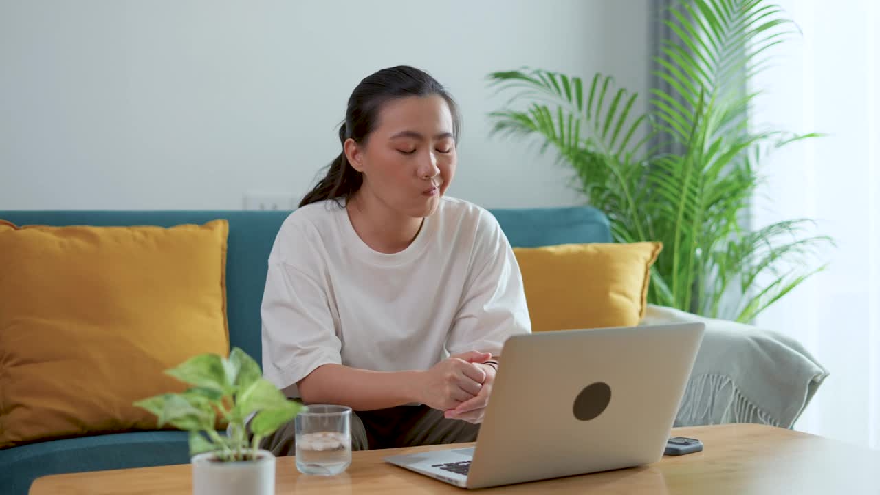 亚洲女人喝着冰水，感觉牙齿敏感，摸着脸颊，坐在客厅的沙发上。视频素材