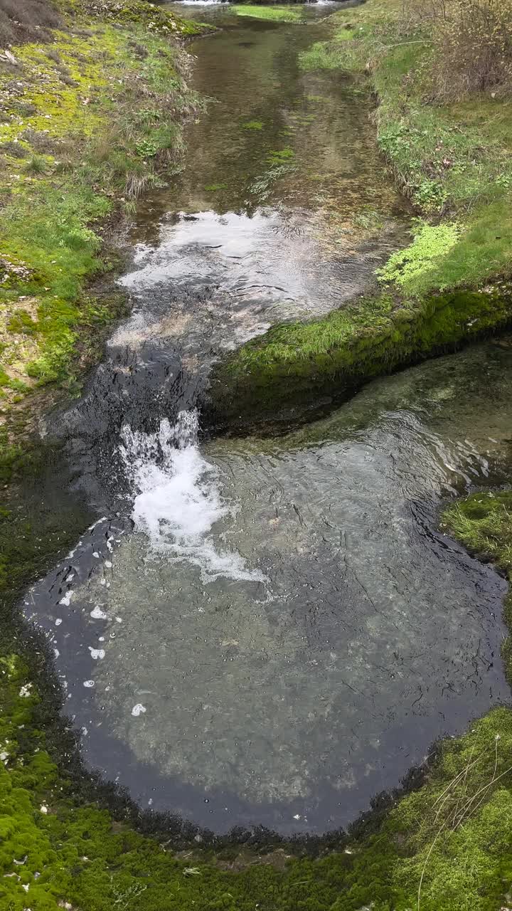 一条清澈清澈的小山河沿着它的河道和绿色的河岸平静地流淌，有两个小瀑布，垂直倾斜，4k视频下载