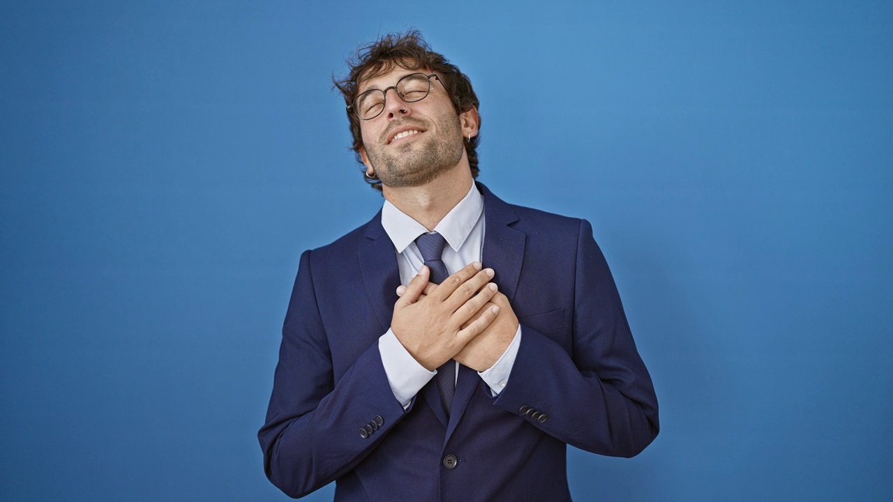 Cheerful young man in business suit, hands on chest, radiates confident gratitude. smiling wide, he embodies health and happiness on a blue isolated background视频素材