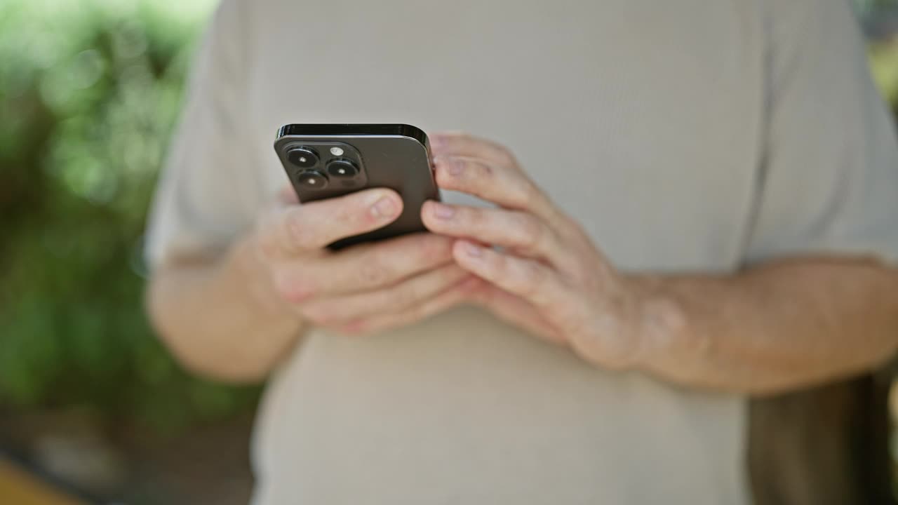 一名穿着休闲衬衫的白人男子在公园外使用智能手机，背景是模糊的绿色植物视频素材