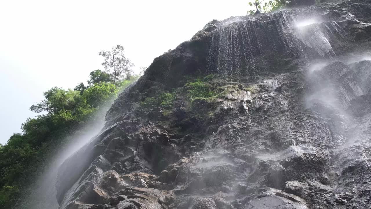 仰望着布满岩石的山顶，细细的瀑布在阳光下流淌视频素材